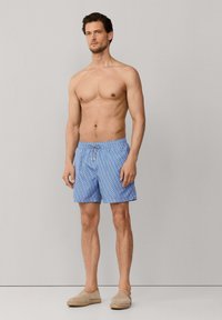 Homme debout pieds nus portant un short de bain bleu à motifs et des espadrilles beige à enfiler sur un fond uni gris clair.