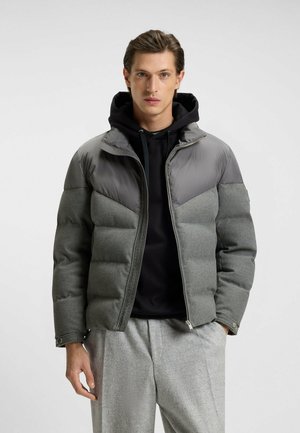 Doudoune grise avec un design de dos en gris plus foncé, dotée d'une fermeture zippée, d'une capuche et d'une texture matelassée ; portée par-dessus un sweat à capuche noir.