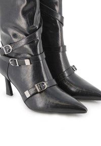 Botas de tobillo negras con punta afilada, hechas de imitación de cuero brillante. Presentan correas decorativas con hebillas en tono plateado y un tacón de 10 centímetros.