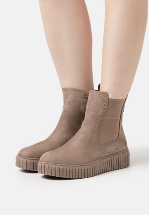 Botas de tobillo de ante topo con suelas de goma gruesas y estriadas, llevadas sobre piernas desnudas sobre un fondo claro y liso.
