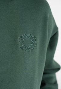 Tela de color verde oscuro con un sutil diseño circular bordado en hilo a juego en la zona del pecho de una prenda.