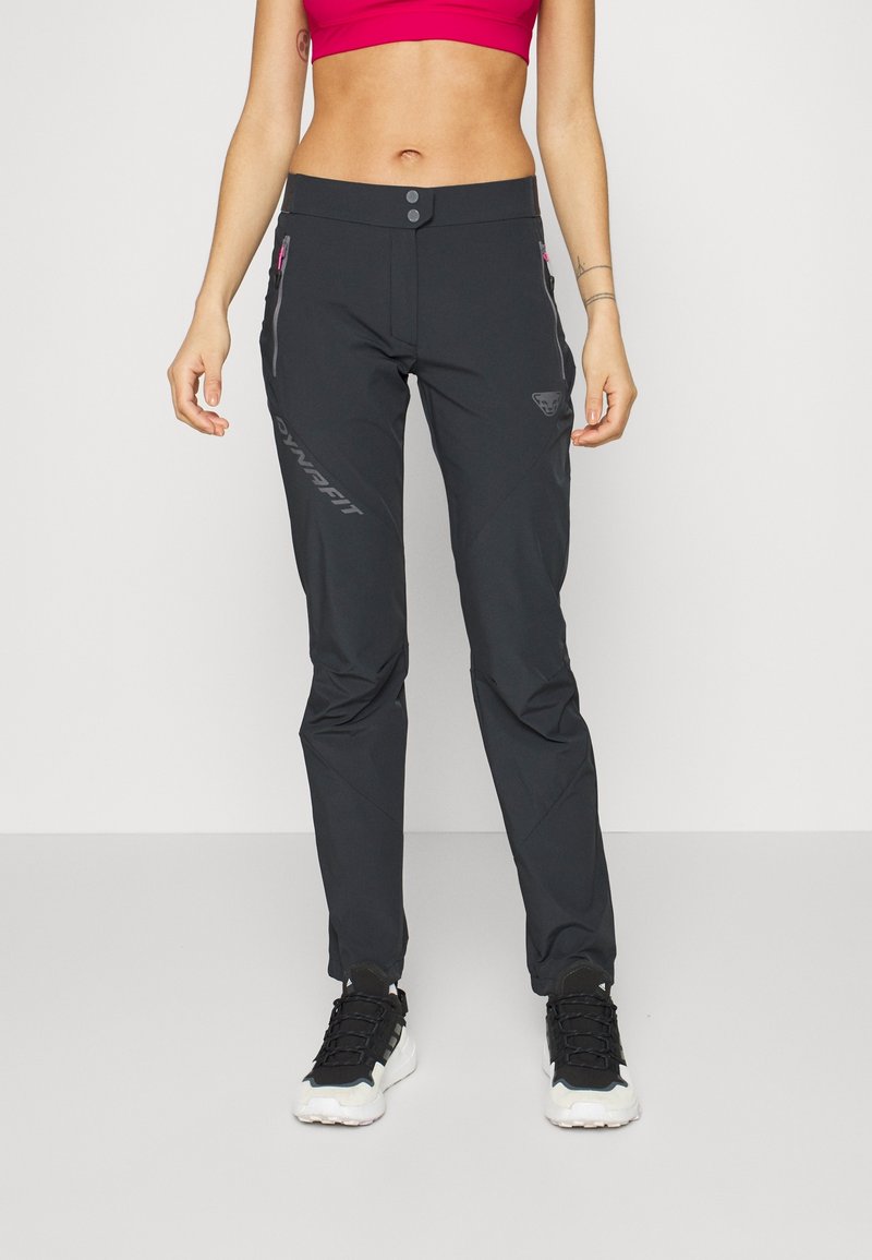 Femme portant un pantalon noir Dynafit avec des poches zippées et des baskets noires et blanches, debout devant un fond uni.