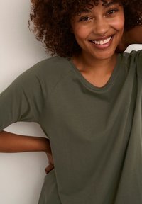 Haut vert olive à manches courtes avec un col rond et une coupe décontractée. Tissu lisse avec un léger éclat, modèle souriant sur un fond clair.