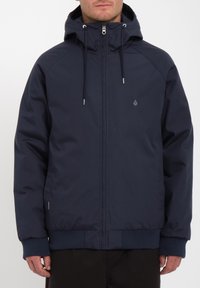 Marineblauwe capuchonjack van gladde stof met een rits, ribgebreide manchetten en zoom. Voorzien van voorzakken en een subtiel logo op de borst.