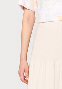 Braccio sinistro e mano di una persona che indossa una maglia a maniche corte con stampa tie-dye in tonalità pastello e una gonna crema con arricciature.