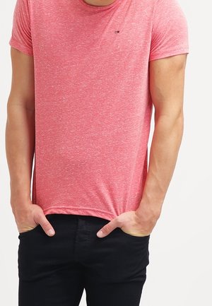 Mand iført en tætsiddende lyserød kortærmet t-shirt og sorte bukser med hænderne i lommerne foran en ensfarvet hvid baggrund.