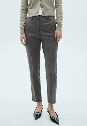 Vrouw draagt grijze op maat gemaakte broek, beige gebreid vest met knopen en zwarte spitse slingback ballerina's, staand tegen een effen achtergrond.