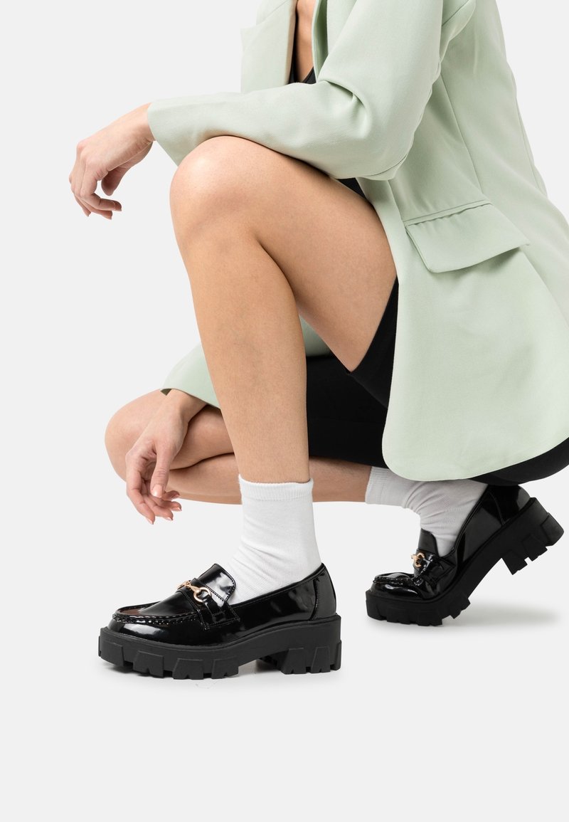 Personne accroupie portant un blazer vert clair, un short noir, des chaussettes blanches et des mocassins en cuir verni noir épais avec un détail de chaîne dorée.