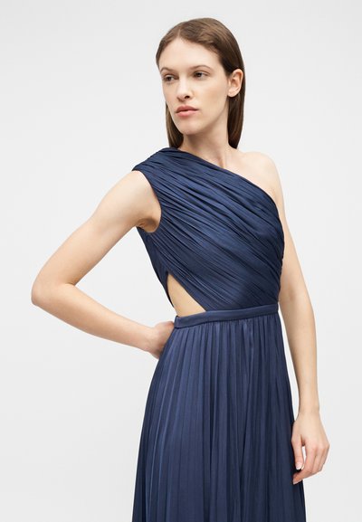 Femme portant une robe plissée bleu marine, une épaule dénudée, sans manches, avec une découpe latérale, posant avec une main dans le dos.