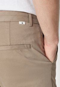 Beige trousers with a smooth texture, featuring a side pocket and a small white label with a symbol. Slim fit design.
