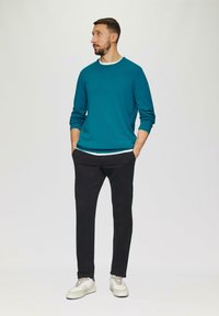Teal langharig vest over een wit shirt, gecombineerd met zwarte broeken en witte sneakers. Eenvoudig, casual ontwerp met een relaxed fit.