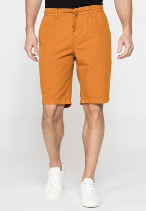Uomo che indossa pantaloncini color senape con orlo risvoltato e vita con coulisse, e sneakers bianche, in piedi davanti a uno sfondo chiaro e uniforme.