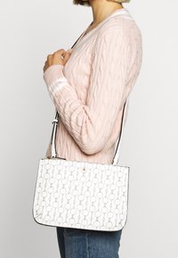 Kvinna som bär en ljusrosa kabelstickad tröja och blå jeans, hållande en vit axelväska med ett cirkulärt länkade mönster.