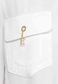 Taschina in tessuto bianco con patta, dotata di occhiello in metallo dorato e una catena con un pendente a forma della lettera "E". Texture liscia.