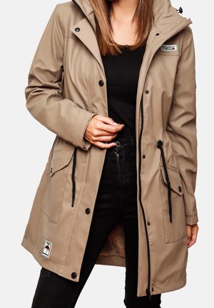 Frau trägt eine beige Kapuzenregenjacke mit Vordertaschen, schwarzen Kordeln und einem schwarzen Hemd sowie einer Hose unter der Jacke.