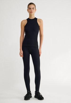 Schwarzes geripptes Tanktop und Leggings aus dehnbarem Material. Verfügt über ein figurbetontes Design und ärmellosen Schnitt, kombiniert mit schwarzen Sportschuhen.