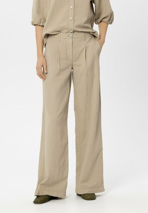 Woman wearing loose beige high-waisted trousers with front pleats, side pockets, paired with a beige buttoned blouse and olive shoes.