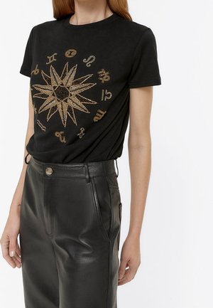 Femme portant un t-shirt noir à manches courtes avec des symboles dorés du zodiaque autour d'un motif étoile, associé à un pantalon en cuir noir à taille haute.