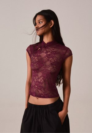 Donna con lunghi capelli scuri indossa un top in pizzo bordeaux trasparente abbinato a pantaloni neri larghi, in piedi con le mani nelle tasche davanti a uno sfondo neutro.