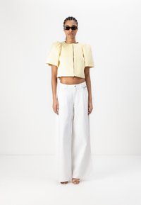 Gelbes, cropped Top mit Puffärmeln und Reißverschluss in der Mitte, kombiniert mit weit geschnittenen weißen Hosen. Das Model trägt dunkle Sonnenbrillen und hat geflochtene Haare.