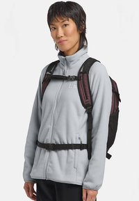 Chaqueta de forro polar gris claro con un cuello alto, usada sobre una mochila granate y negra, que cuenta con correas ajustables y una parte trasera de malla para ventilación.