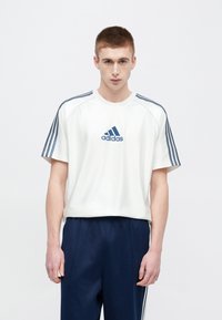 Biele športové tričko s námornícky modrým logom Adidas, s troma námorníckymi pruhmi na rukávoch a zaobleným výstrihom. Dopĺňa ho námornícky modré nohavice.