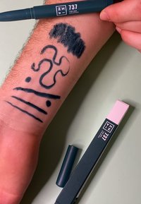 Mørkeblå øyenstift plassert ved siden av en hånd, med streker og mønstre tegnet på huden. Produktet har et slankt design med en matt finish.
