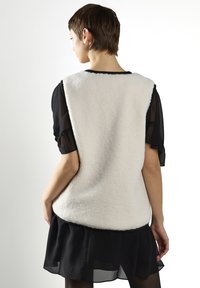 Flauschiges weißes ärmelloses Oberteil mit abgerundetem Saum und schwarzem Abschluss, getragen über einem transparenten schwarzen Shirt, das eine weiche Textur und eine entspannte Passform zeigt.