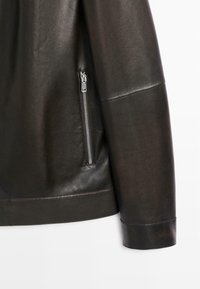 Zwart leren jas met mouw en zijkant waarop zilveren ritsdetails van een zak te zien zijn op glad, gestructureerd materiaal.