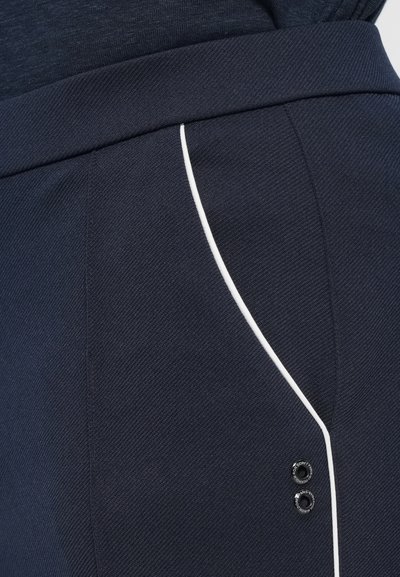 Marineblaue Hose aus strukturiertem Stoff mit weißen Paspeln entlang der Seitentaschen und zwei schwarzen Dekorationsknöpfen in der Nähe des Saums.