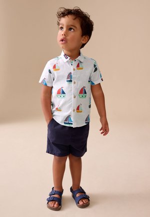 Jongentje draagt een wit shirt met kleurrijke zeilboten, donkerblauwe korte broek en blauwe sandalen, staat met één hand in de zak en kijkt omhoog.