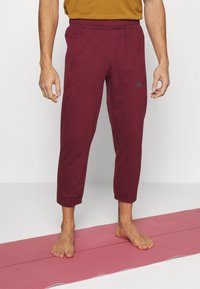 Bordeaux joggings met een elastische tailleband, cuffed enkelontwerp en een klein logoaccent aan de linkerkant. Gedragen op blote voeten.