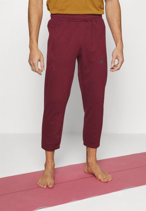 Bordeaux joggings met een elastische tailleband, cuffed enkelontwerp en een klein logoaccent aan de linkerkant. Gedragen op blote voeten.