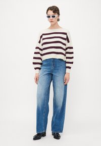 Junge Person trägt einen gestreiften Pullover in Weiß und Dunkelrot, weite blaue Jeans, schwarze Schuhe und eine weiße Sonnenbrille vor einem weißen Hintergrund.