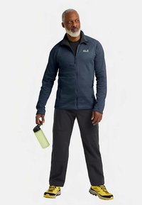 Marineblauwe fleecejas met ritssluiting over een zwart shirt, gecombineerd met donkere broek en gele wandelschoenen. Hand die een groene waterfles vasthoudt.