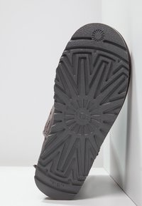 Czarna, teksturowana, gumowa podeszwa z wzorem przypominającym promienie słońca, z logo "UGG" na środku. Częściowo widoczna w profilu na jasnym tle.