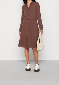 Abito camicia marrone con maniche lunghe, bottoni frontali e cintura da annodare. Abbinato a sandali bianchi con suola spessa e una borsa a spalla color crema.