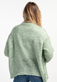 Jasnozielony sweter z dzianiny o luźnym kroju, teksturowanej tkaninie, otwartym splocie i opadłych ramionach. Rękaw jest prosty i nieco dłuższy z tyłu.
