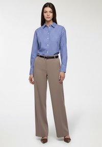 Kobieta stoi ubrana w niebiesko-biały paskowany koszulę zapinaną na guziki, włożoną w szerokie beżowe spodnie z czarnym paskiem oraz brązowe czółenka.