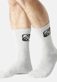 Des jambes portant des chaussettes de taille classique en gris clair côtelé avec un logo géométrique noir sur le côté extérieur, sur fond uni clair.