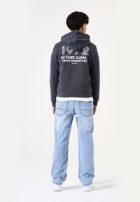 Sudadera gris con capucha y texto blanco en la parte posterior, combinada con jeans holgados de color azul claro y zapatillas blancas. Diseño simple y casual.