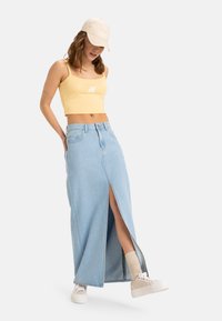 Lyseblå denim maxi-nederdel med en frontslids, høj talje og to sidelommer. Båret med en gul crop-top og en beige kasket.