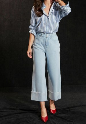 Jean large bleu clair avec ourlet effiloché, associé à une chemise boutonnée à rayures blanches verticales, et des talons rouges pointus.