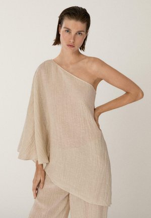 Kvinde med kort brunt hår iført en beige, tekstureret, én-skulders top og matchende løse bukser, stående med hånden på hoften foran en ensfarvet baggrund.