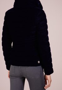 Mörkblå puffjacka med en sammetliknande textur, hög krage och horisontell sömnad. Har en cirkulär logotyp på den nedre vänstra sidan.