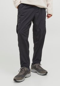 Pantaloni cargo grigio scuro, dotati di multiple tasche, vita elastica e gamba affusolata. Indossati con scarpe sportive grigie, tessuto textured.