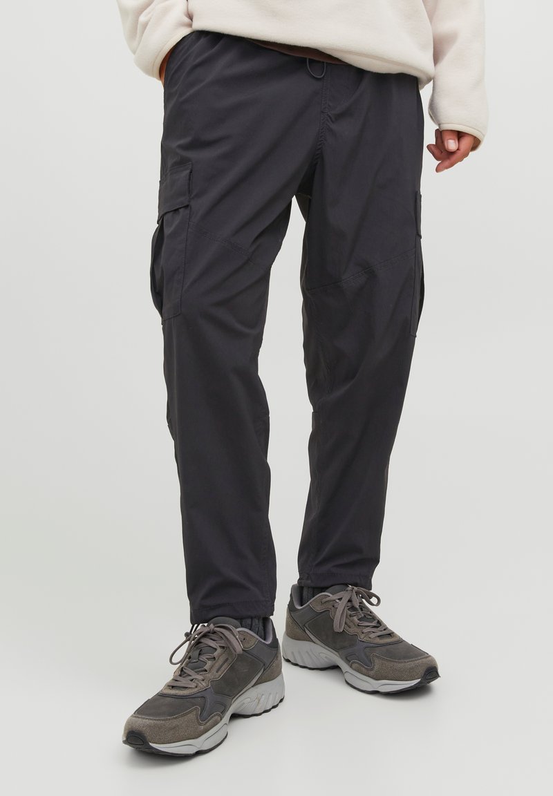 Pantaloni cargo grigio scuro, dotati di multiple tasche, vita elastica e gamba affusolata. Indossati con scarpe sportive grigie, tessuto textured.