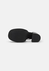 Chaussure à talon haut noire avec une semelle en caoutchouc texturée, un design élégant et un talon proéminent. La semelle présente des rainures horizontales pour l'adhérence.