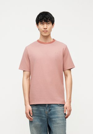 Jeune homme debout, portant un t-shirt rose poudré à manches courtes et un jean bleu, sur un fond blanc uni.