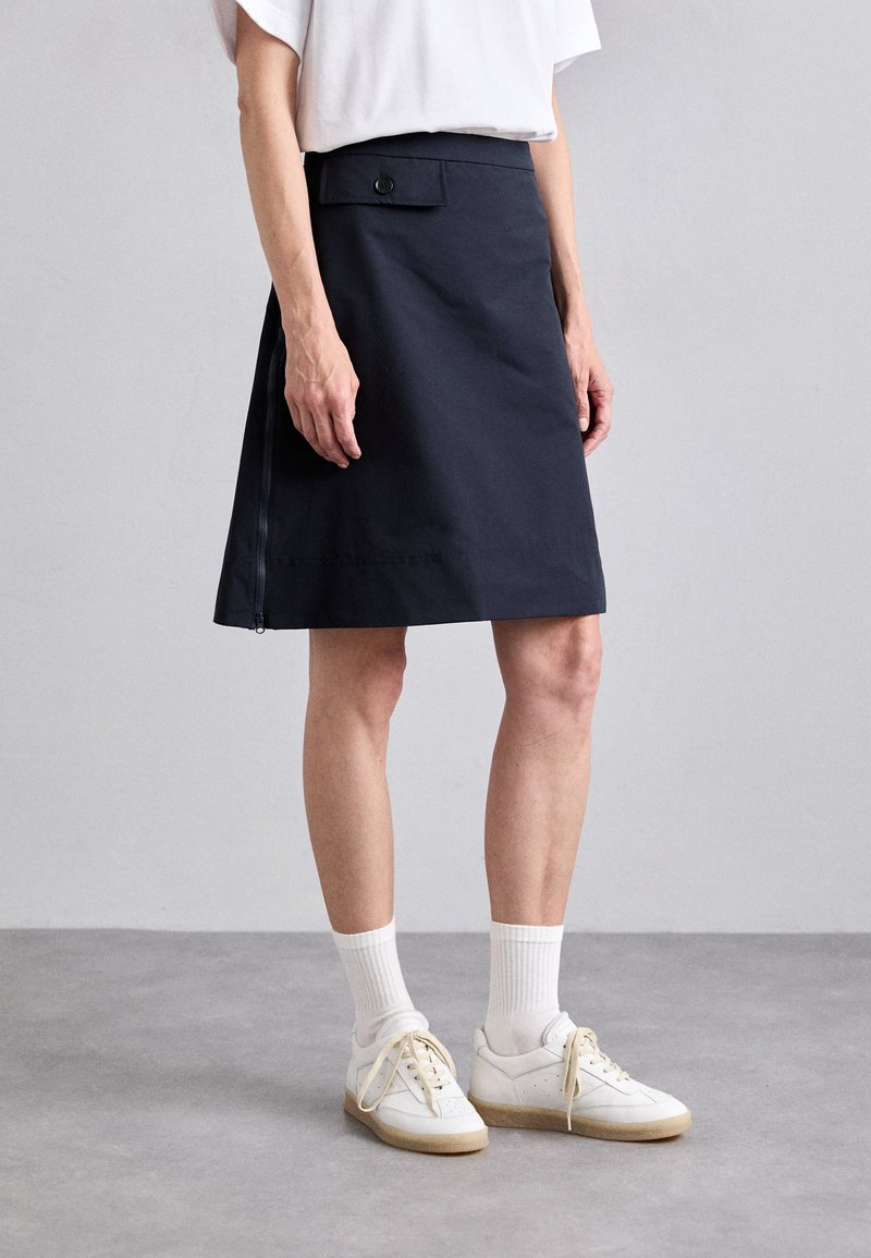 Marineblaue A-Linien-Rock mit verstellbarem Bund, seitlichem Reißverschluss und Knopfdetails. Kombiniert mit weißen Sneakers und strukturierten Socken.
